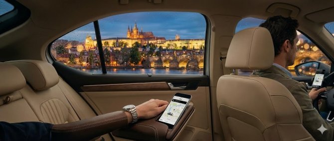 Pohled ze zadního sedadla luxusního vozu během jízdy s Uberem v Praze. Pasažér sleduje aplikaci na telefonu, zatímco řidič řídí. Z deštivého okna je večerní výhled na osvětlený Pražský hrad a Karlův most